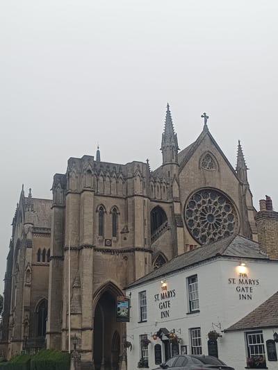 The image captures a view of a large Gothic-style building, likely a church or cathedral, with intricate stone carvings and a prominent rose window. To the right of the larger building, a white, two-story establishment with black lettering that reads "THE ST. MARYS GATE INN" is visible. This inn appears to be a pub or hotel, with signage indicating food and accommodation services. The sky is overcast, suggesting a cloudy or foggy day. The foreground shows some greenery, possibly hedges, and a portion of a dark-colored car. The scene is set in Arun, United Kingdom, as indicated by the context provided.