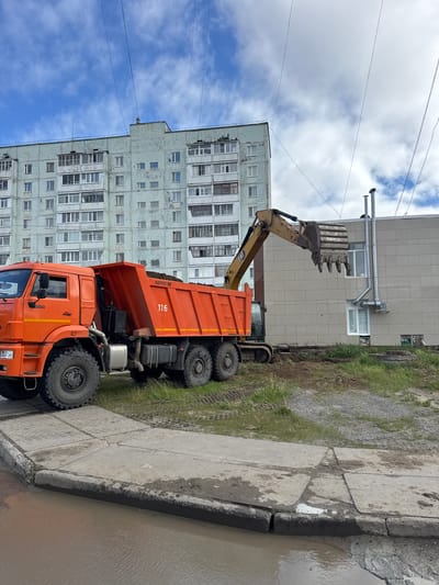 The image depicts a construction scene in Usinsk, Russia, likely during daytime under a cloudy sky. The main subjects are an orange dump truck and an excavator with its bucket raised. The dump truck, with "NEFAZ" and the number "116" visible on its side, appears to be partially loaded or waiting to be loaded with material. The excavator, positioned behind the dump truck, is actively engaged in digging or moving earth, with its large bucket positioned over a trench or an excavation site.

The setting includes a large, multi-story apartment building with a grey facade and numerous windows, characteristic of Soviet-era architecture, and a lighter colored building behind it. Adjacent to the vehicles and buildings is a strip of grass and dirt, indicating ongoing construction or maintenance work. A concrete sidewalk runs along the foreground, with evidence of water pooling, suggesting recent rain or a leak. The overall impression is one of industrial or urban renewal activity. The sky is partly cloudy with patches of blue.