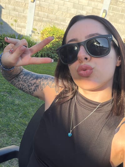 A close-up, front-facing outdoor portrait depicts a female subject in Quilmes Oeste, Argentina. The subject, with medium-length dark hair, wears black rectangular sunglasses, a black sleeveless top, a silver-colored necklace with a blue heart-shaped pendant, and a blue beaded bracelet on her left wrist. She presents a stud piercing on her left nostril and earlobe. Her lips are puckered in a "kiss" expression, featuring matte pinkish-brown lipstick. Her left arm exhibits extensive black ink tattoo work, comprising floral and mandala patterns. Her left hand is raised, displaying a "peace" sign, with fingernails manicured with clear polish, black tips, and a red heart design on the ring finger. The background features a manicured grass lawn, a light-colored brick wall, green foliage, several Agapanthus plants, and a wall-mounted outdoor light fixture, all illuminated by direct sunlight which casts shadows. A portion of a black chair is visible on the lower left.