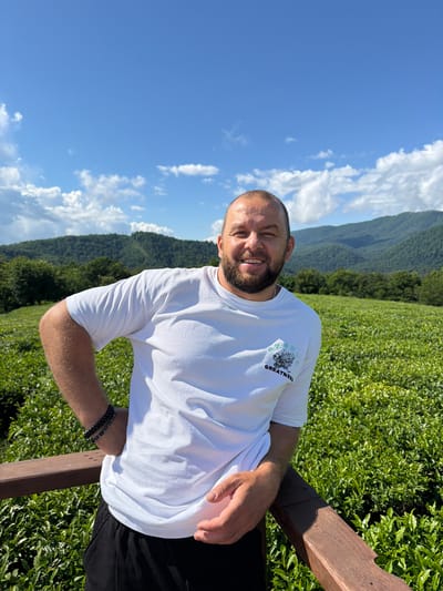 A man with a beard and short hair stands leaning on a wooden railing in front of a lush green tea plantation. He is wearing a white t-shirt with a graphic design and black pants, and two bracelets on his left wrist. The background features rolling hills covered in dense green vegetation, with a bright blue sky dotted with white clouds above. The location is identified as Izmaylovka, Russia.