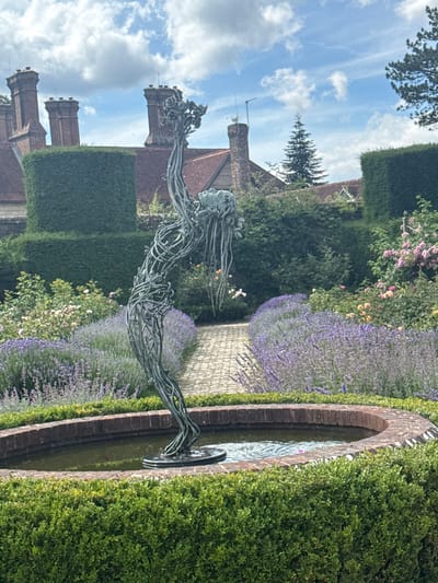The image depicts a garden sculpture of a human figure made of intertwined metal wires, positioned in a circular fountain with a brick surround. The sculpture is centrally located in a well-maintained garden featuring manicured hedges, rows of lavender, and blooming roses in shades of pink and peach. In the background, a stately building with prominent chimneys and red-tiled roofs is visible, suggesting an estate or historical property. The sky above is a mix of blue with scattered white clouds. The scene is set in the city of Mid Sussex, United Kingdom.