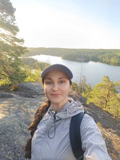 A medium shot selfie shows a woman with brown hair in a braid, wearing a blue baseball cap and a light grey hooded jacket with a dark backpack. She is positioned in the foreground, with her face turned towards the camera. Behind her, a vast natural landscape unfolds, featuring a serene lake surrounded by lush green forests. The setting sun casts a warm, golden glow across the scene, illuminating the trees and the water. The terrain in the foreground is rocky and uneven, characteristic of a natural, elevated viewpoint. The overall scene depicts a moment of peaceful enjoyment of nature. The location is identified as Skriketorp, Sweden.