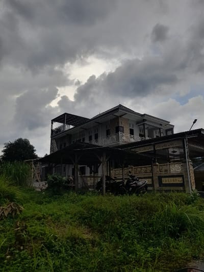 The image displays a multi-story building situated under a heavily overcast sky, indicative of an impending or recently passed rain shower. The photograph is oriented vertically, showing the scene rotated 90 degrees clockwise.
The central subject is a substantial building, possibly a residence or guesthouse, featuring multiple levels and balconies with intricate decorative balustrades. Its architecture incorporates traditional elements, such as ornate carved panels, some of which appear to have unidentifiable script or patterns, particularly visible on the upper section of the facade. The building has a dark-colored roof and appears to be well-constructed.
In the foreground and to the right of the building, there is a dense expanse of lush green grass and assorted vegetation, suggesting an abundant natural environment. Partially visible near the building, likely beneath an overhang or carport, are several motorcycles, indicating human presence, though no individuals are explicitly seen.
The sky is dominated by thick, dark gray clouds, with occasional lighter patches where the sun's diffused light breaks through. This heavy cloud cover creates a soft, subdued lighting condition across the scene. The overall atmosphere is quiet and somewhat muted due to the cloudy weather. The setting suggests a semi-rural or natural area in Kecamatan Prigen, Indonesia, with the local architectural style harmonizing with the verdant surroundings.