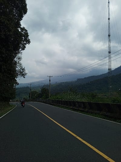 The image depicts a two-way asphalt road, marked by a solid yellow center line, extending from the foreground, curving gently left, and receding into the distance. This scene is located in a mountainous region of Ciloto, Indonesia, under an overcast sky dominated by heavy grey clouds. Dark tree foliage frames the left side of the image. A dark motorcycle, with two visible occupants, travels away from the viewer in the left lane. A dark concrete barrier runs along the right edge of the road, beyond which dense green vegetation covers undulating terrain. Several utility poles, supporting multiple electrical lines, are positioned parallel to the road on the right. In the background, forested mountains are partially obscured by atmospheric haze or low clouds. A tall, grey metal lattice transmission tower stands vertically on a distant forested slope on the right, stabilized by multiple guy wires.