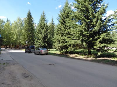 The image depicts a paved road on a sunny day in Ridder, Kazakhstan, with lush green trees lining the right side. On the left side of the road, there is a sidewalk and a dirt patch. Two cars are parked on the road: a dark SUV and a silver sedan. The SUV is closer to the camera and appears to be a Mitsubishi Pajero Sport, with license plate "877 BAX 150". The silver sedan, a Hyundai Solaris, is further down the road with license plate "R096 LCN". In the background, more trees and some buildings can be seen. The sky is blue with a few white clouds. The scene appears to be an urban or suburban street.