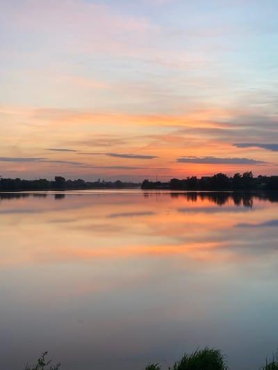 The image depicts a reflection of a sunset sky in a body of water. The sky features streaks of orange, pink, and blue hues. A thin line of trees, potentially on the shoreline, is visible in the middle of the image. Grass is present on the right side of the image, and there are some barely visible structures in the center of the image, which is the town Aramil, Russia.
