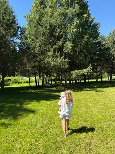 In Копнино, Russia, a bright, sunny outdoor scene displays a wide expanse of vibrant green grass, partially shaded by trees. An adult, viewed from behind with long blonde hair, wears a white t-shirt, white shorts, and dark sandals, carrying a baby. The baby, also seen from behind, wears a white bucket hat and light-colored clothing. Both are walking away from the camera, across the lawn, towards a dense treeline in the background. Strong sunlight creates distinct shadows on the grass. The treeline comprises numerous tall evergreen pine trees interspersed with deciduous trees, including a prominent birch tree with light bark. Within the treeline, a dark rectangular wooden sign is visible hanging from a tree, and a person is relaxing in a dark-colored hammock strung between two trees. A clear, bright blue sky is visible above the trees.