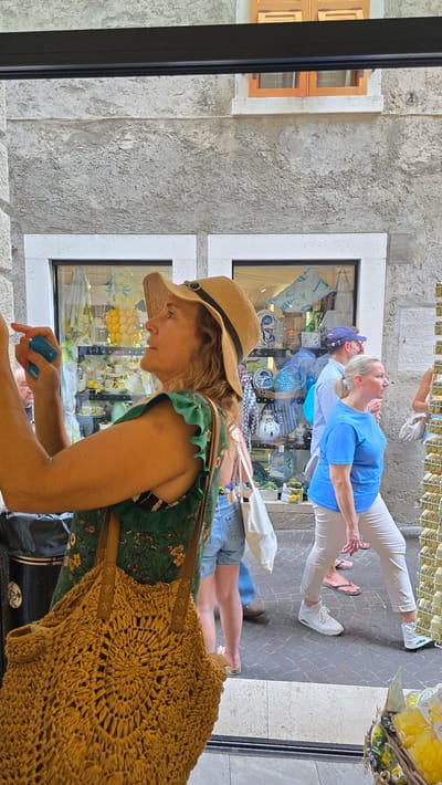 The image shows a woman in the foreground wearing a straw hat and a green floral dress. She is leaning forward, looking at a blue credit card she holds in her hand. Behind her, through a storefront window, a display of various items, possibly souvenirs or local products, is visible. Further in the background, two people are walking away from the viewer. The woman in the blue top is wearing white pants and white sneakers, while the person behind her is wearing a blue and white striped shirt and a blue baseball cap. The scene appears to be an outdoor street market or shopping area, possibly in a tourist location given the souvenirs in the window. The architecture suggests a European setting. The lighting indicates it is daytime, and the clothing suggests warm weather. The overall mood is one of casual shopping. The credit card in the foreground has "Visa" and "digital" written on it.