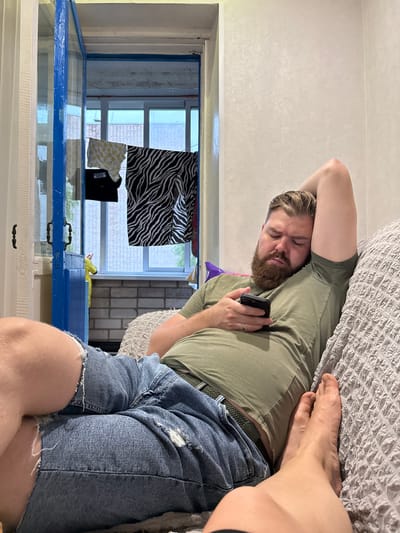 A man is lying on a textured gray couch, holding a black smartphone in his right hand. He is wearing a faded olive green t-shirt and ripped denim shorts. His eyes are closed, and his head is tilted back, resting on his right arm which is bent behind his head. A thick brown beard covers his chin and jawline. The couch is positioned in an indoor setting, with a partially open blue door visible to the left of the frame. Beyond the door, laundry is hanging on a line, with various items including a yellow patterned garment and a black and white striped fabric. The background appears to be a balcony or porch area with windows and brickwork. The flooring visible through the doorway is tiled. The overall lighting suggests an indoor environment, possibly during the daytime given the light coming through the windows.