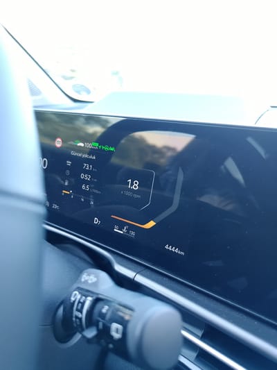 A car's digital dashboard displays a journey log. The log shows a current speed of 100 km/h, a daily distance of 73.1 km, and an average fuel consumption of 6.5 L/100km. The engine is running at 1000 rpm, indicated by "1.8 x 1000 rpm". The current outside temperature is 23°C. The car is in drive gear "D7" and the temperature gauge shows a reading between 50°C and 130°C. The total mileage of the car is 4444 km. The dashboard is illuminated by sunlight. A windshield wiper control stalk is visible in the foreground. The image does not provide information about the location in Garbsen, Germany.