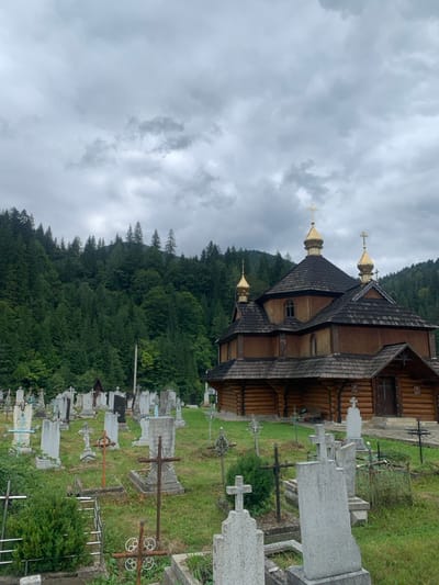 The image displays a static scene of a graveyard and a wooden church, located in Tatariv, Ukraine, under an overcast sky. A prominent wooden Orthodox church, constructed from horizontal logs with a dark shingled roof, stands in the midground to the right. It features three golden, onion-shaped domes, each topped with a cross. The foreground and left midground are filled with a densely packed graveyard, containing numerous gravestones and various types of crosses made of stone and metal, set within an unkempt grassy area. Behind the church and cemetery, a steep hillside is covered by a dense forest of dark green coniferous trees. The sky above is entirely obscured by thick, grey clouds. No individuals are present in the scene.