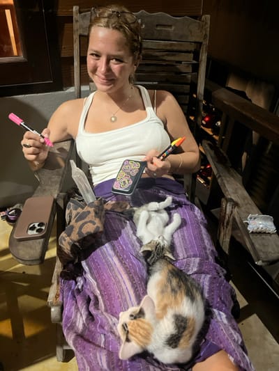 A light-skinned female, appearing to be between 20 and 35 years old, is seated in a wooden slatted chair in Baan Nai Wok, Thailand. She has light brown hair pulled back, wears a white tank top, and a gold-colored sun pendant necklace. She smiles directly at the camera. In her left hand, she holds a pink Posca marker, and in her right hand, she holds a multi-colored (yellow, orange, black) Posca marker along with a black object featuring vibrant yellow, pink, and green swirling patterns.

Two kittens are resting on a purple patterned fabric draped across her lap/legs. One is a calico kitten (white, orange, black) lying on its stomach, facing away from the camera. The other is a lighter-colored kitten (possibly tabby or white/grey) lying on its back with its paws extended upwards, also facing away.

To the left, a brown-cased smartphone (appearing to be an iPhone Pro model) rests on the chair's armrest. A leopard-print pouch is positioned on the fabric, containing a translucent, cylindrical object identified as a clear glass or plastic chillum/pipe. On the right armrest, a small white ceramic dish shaped like a cat is visible. The immediate background shows dark wooden paneling, with red objects partially visible in a rack-like structure to the right. The scene is illuminated by artificial light.