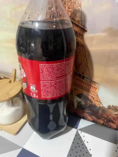 A close-up, eye-level shot shows a plastic bottle of Coca-Cola sitting on a patterned surface. The bottle is mostly dark, with a red label that has white lettering and a white Coca-Cola logo. The label contains information in Russian. To the left of the bottle, a sugar bowl filled with white sugar is visible. In the background, a large image of the Eiffel Tower is displayed. The lighting is artificial and casts shadows. There are no people in the image, and no actions are taking place. The image was taken in the city of Панфилово, Kazakhstan.