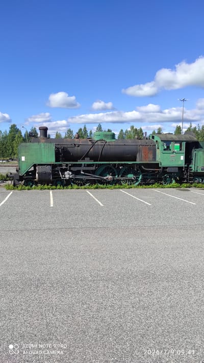 An old, green and black steam locomotive sits on tracks in a paved area in Otanmäki, Finland. The locomotive is partially covered in green paint, with areas of rust visible on the boiler. It has large, spoked wheels and a visible cabin. The background consists of a line of trees under a blue sky with scattered white clouds. The foreground is a paved surface marked with white lines, resembling a parking lot. There are no people visible in the image.