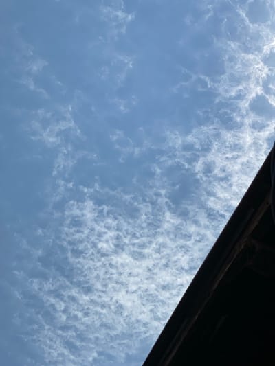 The image depicts a sky filled with scattered, wispy clouds, predominantly white and light grey, against a clear blue background. The bottom right portion of the image is occupied by a dark, angled structure, possibly a rooftop or overhang, with visible wooden elements and dark shadowing. The composition suggests an outdoor scene, likely taken from ground level or a lower vantage point, looking upwards. The clouds exhibit a textured pattern, with some areas appearing denser and others more translucent, indicating varying levels of atmospheric moisture. The lighting suggests a daytime scene, possibly late morning or early afternoon, with the sun filtering through the clouds and creating subtle highlights. The overall impression is one of an ordinary sky on a partly cloudy day, with an architectural element framing the view. There are no people or discernible activity in the image. The image does not contain enough information to determine its specific location within Banjarmasin, Indonesia.