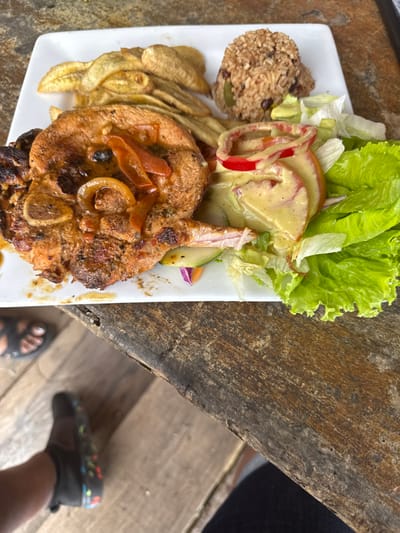 A meal is displayed on a white rectangular plate, centrally positioned on a rustic, weathered wooden table. The plate contains a grilled pork chop topped with sautéed red bell pepper strips and onion rings in a light brown sauce, exhibiting some charring. Adjacent to the meat are golden-brown fried plantain slices and a scoop of rice mixed with black beans and green vegetable pieces. The plate also features a salad composed of green leaf lettuce, sliced red tomatoes, red onion rings, and sliced cucumber, all covered in a creamy, light yellow-green dressing. The wooden table surface shows pronounced grain, cracks, and wear. Below the table, a foot wearing a dark sandal with a thick strap is visible on the left, and another foot wearing a dark water shoe with multi-colored speckled soles is visible towards the bottom-center. This scene is situated in the city of La Ceiba, Honduras.