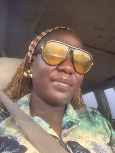 A woman with dark skin and light brown box braids is seated inside a vehicle, likely in Elelenwo, Nigeria. She is wearing a patterned headband, a button-up shirt with a green, yellow, and black abstract or tropical print, and a light brown seatbelt across her chest. Large, dark-framed aviator-style sunglasses with reflective yellow lenses obscure her eyes; reflections in the lenses show an outdoor environment with structures and light. A glossy substance is applied to her lips, and a large, gold-toned, C-shaped earring is visible on her left ear. The vehicle's headliner, appearing dark grey, is visible above her, and a car window is visible to her right, showing a hazy bright sky or distant view.