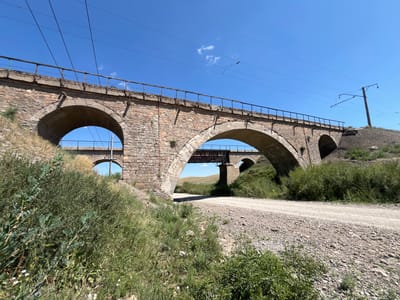 The image shows a dry, dusty rural road passing under a large, old stone arched railway bridge. The bridge is made of rough-hewn stones and appears to be in disrepair in some areas, with visible concrete patches and rusty metal railings. The road is unpaved and covered with gravel and dirt. On either side of the road, there is dry grass and green bushes, suggesting a semi-arid environment. In the background, rolling hills are visible under a clear blue sky. Another, smaller bridge or viaduct can be seen further in the distance, also made of stone arches and supporting what appears to be a railway track with overhead electrical lines. The context provided places the scene in Astakhovka, Kazakhstan. There are no people or animals visible in the image. The overall impression is one of a remote, somewhat neglected infrastructure in a natural, open landscape.