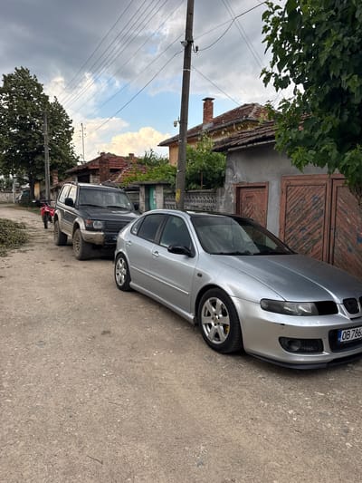 Two cars are parked on a dirt road in Bratya Daskalovi, Bulgaria, next to a residential building with wooden doors and a red-tiled roof. A silver sedan is parked in the foreground, facing right, with its front bumper and headlights visible. Behind it, a dark-colored SUV is parked, facing left, with its front end slightly visible. The SUV has a license plate with the letters "BG." A red motorcycle is parked to the left of the SUV. The scene is surrounded by trees and power lines crisscross the sky. The ground is covered in dirt and small stones.