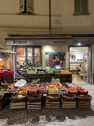 This image captures an outdoor street view of a bustling fruit and vegetable shop, likely in Sondrio, Italy, given the provided context. The time of day appears to be evening or night, indicated by the artificial lighting illuminating the storefront and produce displays, casting shadows on the cobblestone pavement.
The shop, identified by the signs "FRUTTA" and "BUNDIN" above its entrance, features an extensive array of fresh produce displayed on wooden crates and wicker baskets on the sidewalk. Visible fruits include oranges, apples, lemons, kiwis, pineapples, grapes, plums, and what appear to be mandarins or clementines. Vegetables like lettuce and cabbage are also present. A decorative wooden wagon or cart is used as part of the display.
The shop's interior is visible through large glass doors and windows, revealing more shelves stocked with items. A prominent poster inside a window shows a person on a moped with "BUNDIN" and "FRUTTA" text.
To the left of the shop, a building facade with closed green shutters and a visible street sign adds to the urban setting. The street sign clearly reads "PIAZZA QUADRIVIO", providing a specific location cue within the town. The ground is paved with cobblestones.
No people are visible in the scene, which focuses entirely on the architecture and the vibrant display of fresh produce. The overall atmosphere suggests a well-stocked and inviting local greengrocer.