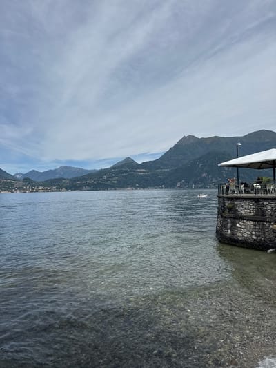 The image depicts a serene lakeside scene in Gittana, Italy. In the foreground, clear water laps against a pebble-strewn shore. The mid-ground is dominated by a calm lake, reflecting the overcast sky. In the background, a range of lush green mountains rise, dotted with small villages. On the right side of the frame, a stone-built structure juts out into the water, topped by a patio with tables and chairs. A white umbrella provides shade for a few patrons enjoying the view. A small boat is visible in the distance on the lake. The overall atmosphere is peaceful and picturesque.