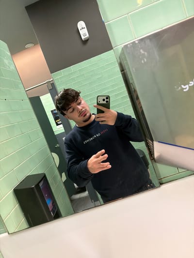 The image depicts a male individual taking a mirror selfie inside what appears to be a public restroom. The subject, seen from the chest up, has dark brown hair, a mustache, and a goatee. He is wearing a dark blue long-sleeved sweatshirt with the reversed text "LION OF PORCHES" visible on the chest. He holds a dark green smartphone in his right hand to capture the image, while his left hand is open with fingers slightly splayed.
The restroom environment features light green rectangular tiles covering the walls. Reflected in the mirror are a metallic silver hand dryer on the right, displaying some residue marks, and a black rectangular soap dispenser with a translucent window showing blue liquid on the left. A small white electronic device is mounted on a dark gray wall section in the upper background. A dark gray door with a horizontal metal handle and a partially visible poster are also reflected in the background. A white countertop forms the base of the mirror in the foreground. The ceiling appears light gray with a recessed circular light fixture. No specific visual information within the image identifies the location as Penafiel, Portugal.