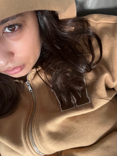 A close-up image features a person with dark brown hair lying down, with their face partially visible. The focus is on their eye, nose, and lips. They are wearing a beige zip-up hoodie, with the zipper partially open. The hoodie features a dark brown patch with light-colored stitching. The texture of the fabric is visible, and the overall lighting creates shadows and highlights. The background appears to be a dark-colored surface.