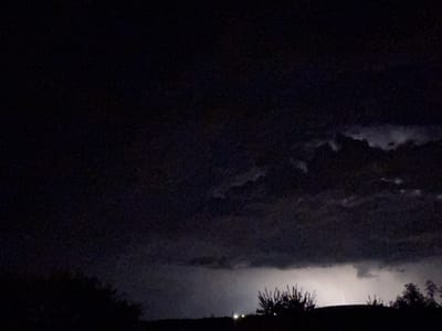 A nighttime image captures a distant lightning strike illuminating dark, stormy clouds above a silhouetted landscape. The foreground shows the dark outlines of trees and vegetation against the horizon. The scene appears to be an outdoor, rural or semi-rural environment, with no identifiable structures or people visible. The lightning flash is the brightest element in the image, suggesting a powerful thunderstorm. The overall mood is dramatic and somewhat ominous due to the darkness and the electrical storm. There are no specific landmarks in Mlada Gvardia, Bulgaria, visible in this image.
