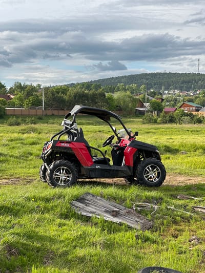 A red and black all-terrain vehicle (ATV) is parked on a dirt and grass path in the foreground. The ATV has a roll cage, a steering wheel, and large tires. In the background, there is a grassy field with some weathered wooden planks on the ground. Beyond the field, there is a village with houses and trees, and a forested hill with a radio tower on top. The sky is cloudy with some patches of blue. The scene appears to be set in Bolshaya Rechka, Russia.