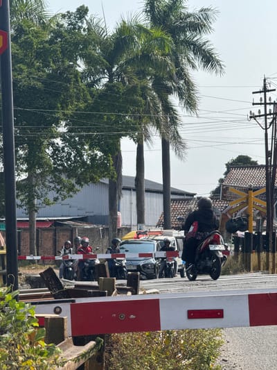 This media file depicts a street scene in Kebulusan, Indonesia, likely during daytime with clear weather, suggesting a warm climate. The image is vertically oriented and features a railway crossing with a red and white barrier arm. Several individuals are present, primarily on motorcycles. One person, wearing a black jacket and helmet, is on a motorcycle, facing away from the camera. Behind them, other motorcycles are visible, some with riders.

The background reveals elements of Indonesian architecture, including buildings with tiled roofs and corrugated metal siding. Palm trees and other lush green vegetation are abundant, characteristic of tropical environments. Utility poles with numerous wires are a prominent feature, suggesting the presence of electricity and telecommunication infrastructure.

The scene captures a moment of waiting or passing through a barrier, possibly at a railway crossing. The overall atmosphere appears to be one of everyday life and transit. There is no explicit indication of any specific event, but the presence of multiple vehicles and people suggests a moderately busy area.

A visible license plate on one of the motorcycles reads "AA 3936 ZIH". The red and white barrier at the crossing, along with the general appearance of the surroundings, strongly points to a location in Indonesia. The lighting suggests it is not early morning or late evening, but rather midday.