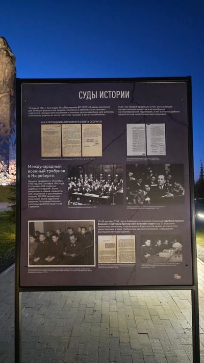Here is a description of the image:
**Scene:** The image shows an informational display panel, possibly part of an outdoor exhibit. The panel is black with white and black text and historical black and white photographs. The panel’s subject matter appears to be about war crimes trials, specifically the Nuremberg trials and trials in Khabarovsk, Russia.
**Text (translated from Russian):** The main title is "COURTS OF HISTORY”. The text below the heading references a decree from April 1943 concerning punishment for Nazi-Fascist criminals, spies, and traitors. It further describes this decree as a foundational legal act for trials against Nazi criminals. The panel also mentions the International Military Tribunal in Nuremberg, and that the trials occurred from November 20, 1945, to October 1, 1946. It states that there were 403 open court sessions with 360 witnesses examined. The panel indicates the trials of former members of the Kwantung Army in Khabarovsk in December 1949.
**Images:** The images on the panel include photographs of legal documents, images of the Nuremberg trials, and photographs of individuals accused of war crimes during the Khabarovsk trials.
**Environment:** The panel is outdoors, with a dark blue sky in the background. There is a stone statue or monument and some greenery visible below the display. The ground nearby is paved.
Based on the identified location in the city of Zaitsevo (Verevskoe S/P), Russia, this display may be a local exhibit or memorial related to World War II and its aftermath.
