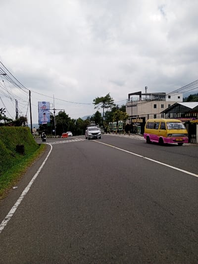 The scene depicts a multi-lane asphalt road in Ciloto, Indonesia, under an overcast sky. The road extends from the foreground into the mid-ground, flanked by commercial and natural elements. On the immediate right, a yellow and pink public transport van (Angkot) with license plate F 7982 TB is positioned facing the viewer, partially on the road's shoulder. Adjacent to the van is a commercial establishment featuring signage for "Nemu Kopiko," comprising a multi-story building with a flat roof and balcony, and an adjoining single-story structure with a dark roof and large windows.
On the left side of the road, a grassy embankment borders the asphalt. Further along the left, multiple utility poles support a dense network of electrical and communication wires. A large billboard displaying a male political figure in a white shirt and peci cap is prominently visible. In the middle distance on the road, a silver SUV equipped with a roof rack is traveling away from the viewer in the left lane. Behind this SUV, a white sedan is observed turning right from a side road, partially obstructed by orange traffic barriers. Further left, a person on a white scooter, wearing a white helmet and dark attire, is positioned at an intersection, appearing to prepare for a left turn onto the main thoroughfare. Green foliage, trees, and distant mountainous terrain complete the background.