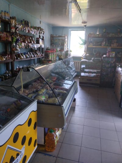 The image depicts the interior of a small grocery store, likely in Novoselivka, Ukraine. The scene is dominated by shelves stocked with various food items and a refrigerated display case filled with products. Sunlight streams in from a window at the far end of the store, suggesting it is daytime.

The shelves are laden with packaged goods, including bottles and bags of what appear to be snacks and dry goods. The refrigerated display case, a prominent feature, contains an assortment of items in clear plastic packaging, likely processed meats or other perishable food products. To the left, a freezer chest with a distinctive yellow and black polka dot design is partially visible. A large plastic bottle of amber-colored liquid, possibly oil or juice, sits on the floor near the display case.

The store appears to be sparsely stocked, and the lighting is somewhat dim, with a bright area near the window. The floor is tiled in a light color. There is no activity or people visible in the image, giving it a still-life quality.

There is some visible text on the yellow freezer chest, which is partially obscured but appears to include the word "MEAT". The overall impression is of a functional, no-frills local shop.