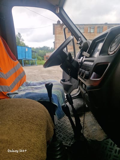 The image captures the interior of a truck cab, likely parked outdoors. The foreground shows a brown towel draped over a seat, with a blue and white striped towel covering another part of the seat. A black gear shifter is prominent. To the left, an orange safety vest with reflective stripes is draped over the seat.
The interior dashboard is visible, with a steering wheel, various buttons, and a car mount for a phone. The perspective suggests the photo was taken from the passenger seat, looking towards the driver's side and out the front window.
Outside the cab, a blue metal structure with stairs is visible, along with trees and a brick building in the background. The ground appears to be a gravel or dirt lot. The sky is overcast.
The text "Galaxy S22+" is watermarked in the bottom left corner, indicating the device used for the photograph. The scene is likely located in Zheleznogorsk-Ilimsky, Russia, based on the provided context.