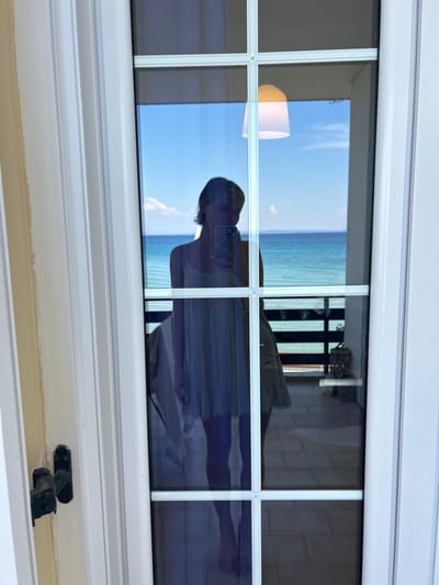 This image shows a reflection in a window. The reflection appears to be of a person wearing a hat and a light-colored dress, possibly standing on a beach or near a body of water. The person is looking down, and their face is not clearly visible. In the reflection, there is a clear blue sky with some clouds and a vast expanse of blue water, suggesting a sunny day at the coast.

Inside the room, partially visible through the window, is a lampshade and some decorative items, including what looks like a woven basket. The window itself is divided into multiple panes by white frames. The lower part of the image shows the exterior of the window frame with a latch.

The overall impression is one of a relaxed, possibly vacation setting, given the beach-like scenery reflected in the glass and the casual attire of the person. The bright sky and clear water suggest it is daytime, likely late morning or afternoon. The location context places this scene in Arkadii Municipal Unit, Greece, which is consistent with the coastal imagery. There is no visible text in the image.