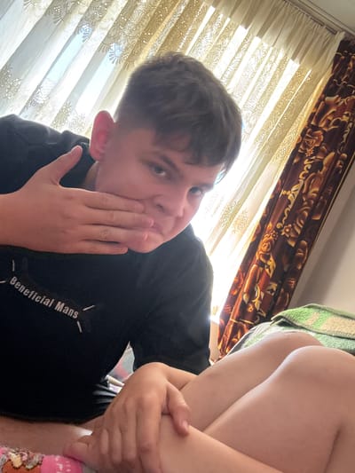The image is a selfie of a person with short, dark hair. They are wearing a black t-shirt with the words "Beneficial Mans" printed on it. The person is holding their hand up to their face. The background consists of curtains and a patterned throw. It is unknown if this image was taken in Damianitsa, Bulgaria.