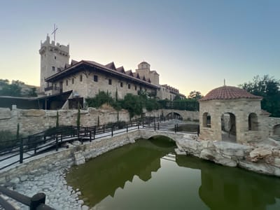 The image depicts a stone structure resembling a medieval castle or monastery, set against a clear, pale sky characteristic of dawn or dusk. A large, multi-story building with a prominent tower topped by a cross dominates the background. This main structure has a long, sloping roof with numerous gables and is constructed from light-colored stone. In the foreground, a tranquil, greenish body of water is bordered by a stone wall and a paved walkway with a black railing. A small, arched stone bridge crosses the water, connecting the walkway to another part of the complex. To the right of the water is a small, circular stone structure with a tiled, domed roof, also topped with a cross. The overall scene suggests a peaceful, possibly historical, or religious site. The lighting indicates the early morning or late evening, with soft, diffused light casting gentle reflections on the water. No people or text are visible. The presence of stone construction, architectural style, and the cross atop the structures are consistent with historical religious or defensive buildings. The scene evokes a sense of serenity and historical gravitas.