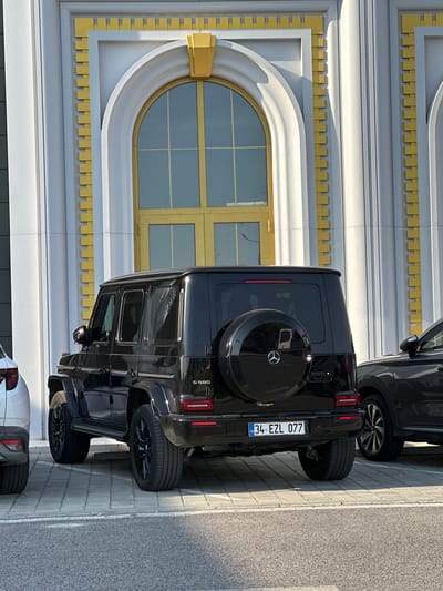 The image captures the rear three-quarter view of a black Mercedes-Benz G-Wagen, model G 580, parked on light grey pavers in front of a distinct architectural building. The vehicle displays a Turkish license plate, "34 EZL 077", indicating its registration in Istanbul, Turkey. A black spare tire cover, bearing the Mercedes-Benz logo, is mounted on the rear door. Black multi-spoke alloy wheels are visible on both the front and rear axles. To the left, a portion of a white SUV is visible, showing its rear quarter panel. To the right, part of a dark-colored SUV is partially visible, showing its front wheel and side.
The background features a building with a light grey or off-white facade, accentuated by prominent gold/yellow decorative elements. A large, arched window or doorway with a golden frame and internal mullions is centered behind the G-Wagen. The building's architecture includes vertical fluted pilasters and decorative rectangular gold blocks arranged in a repetitive, stacked pattern along vertical edges. Arched architectural details above the windows/doorways also feature golden accents. The scene is illuminated by strong, natural sunlight, casting defined shadows. In the immediate foreground, an asphalt road surface is visible, separated from the paved parking area by a thin white line. No individuals are present in the image. The specified location of Kapitan Andreevo, Bulgaria, is consistent with the presence of a Turkish-registered vehicle, given the town's position at the Bulgarian-Turkish border.