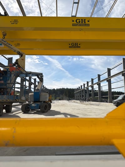 In Janowice, Poland, a construction site is captured under a partly cloudy sky. The foreground features a bright yellow overhead crane beam, obscuring the lower portion of the image. Two similar yellow beams run horizontally across the midground, each bearing the "GH" logo and the website "www.ghcranes.com". Below the top beam, the text "CE 2024", "N° 128893", and "GNP A5" is visible. The lower yellow beam also displays "CE 2024", "N° 128894", and "GNP A5".
To the left, a blue aerial work platform with the "JLG LIFT" logo is positioned. Next to it, another blue aerial work platform, this one a Genie brand, is visible with "x4" marked on its side. In the background, the skeletal structure of a large building is under construction, with numerous steel beams erected. A dark-colored van is parked on the right side of the construction area. The ground is paved, and a line of trees forms the treeline in the distance.