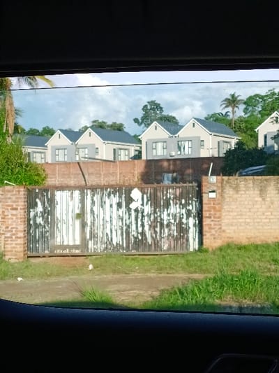 The image captures a residential street scene in Harare, Zimbabwe, viewed from inside a vehicle. A large, weathered black metal gate with extensive peeling white paint dominates the midground, flanked by brick pillars and a low brick wall to the right. A dirt path leads to the gate, bordered by green grass. A house number "13" is affixed to the right brick wall. Beyond the gate and a dividing brick wall, multiple modern, single-story houses with light grey facades and dark grey gabled roofs are visible. These houses feature numerous windows and some visible external air conditioning units. The background consists of lush green trees and foliage, including several palm trees, beneath a clear blue sky with scattered white clouds. No individuals or dynamic actions are present.