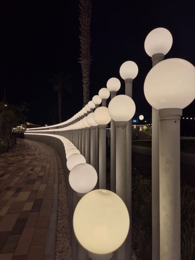 An outdoor perspective shot shows a pathway lined with spherical, illuminated lights in Fujairah, United Arab Emirates. The path is paved with rectangular bricks in a checkerboard pattern of tan and dark brown colors. To the right of the path, a series of evenly spaced, cylindrical posts topped with white, spherical lights extend into the distance, forming a curved line. The lights are illuminated, casting a soft glow on the surroundings. In the background, silhouetted palm trees are visible against the dark night sky. Further in the distance, there are some blurred structures with lights, possibly buildings or an establishment. The overall scene depicts a well-lit walkway at night, likely a public area or promenade.