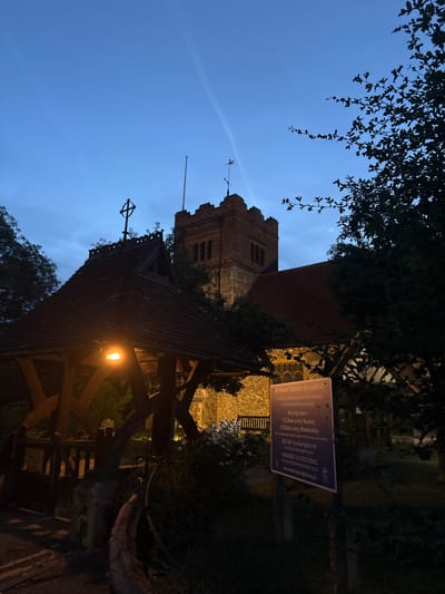 The image shows a church in the city of Chelmsford, United Kingdom, at dusk. The church is made of brick and has a tall tower. In the foreground, there is a wooden structure with a red tiled roof, possibly a lychgate, with a light on. A signpost to the right of the lychgate displays information about "All Saints' Church, Springfield". The sky is a deep blue with some clouds and a faint contrail. Trees with dark leaves are visible on the right side of the image, silhouetted against the sky.