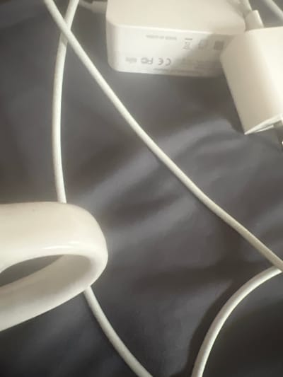 The image displays a close-up, overhead view of white electronic cables and a white power adapter on a dark gray, textured surface, possibly fabric or a similar material. The power adapter is rectangular and has some markings and symbols on it, including "CE FC". The cables appear to be intertwined and are mostly out of focus in the foreground, with the power adapter more in focus in the background. The lighting casts shadows on the surface, creating a dim and somewhat indistinct environment. There are no people visible in the image. The location of West Hartford, United States of America, is not visually determinable from the image content.