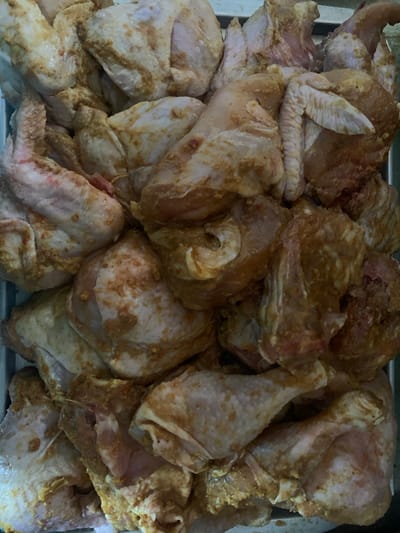 A close-up, overhead shot displays a large quantity of raw chicken pieces, cut into various sections such as wings, drumsticks, and thighs. The chicken is coated in a marinade or spice rub that appears to be a mixture of light brown and yellow hues, with some red flecks, suggesting the presence of turmeric, chili, and other spices. The chicken pieces are densely packed in a metal tray or container, filling the entire frame. The lighting is even, casting minimal shadows, and the focus is sharp, highlighting the texture of the raw meat and the seasoning. The scene is devoid of any human presence or specific environmental details that would indicate a location within the city of Fajo 2 Division Range, Nigeria, or any other specific geographical context.