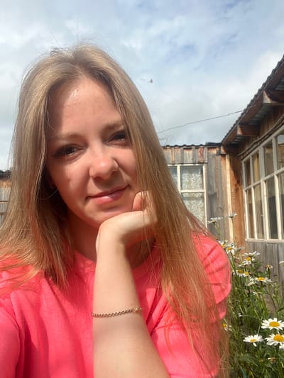 A young blonde woman with fair skin is pictured from the chest up, taking a selfie outdoors. She is wearing a bright pink crewneck shirt and a delicate gold bracelet on her right wrist. Her left hand rests on her chin, and she is smiling slightly. Her long blonde hair falls around her shoulders. The background suggests a rural or suburban setting, with a rustic wooden building and a patch of white daisies. The sky is partly cloudy. The image appears to be taken on a sunny day. The context of Kovriga, Russia, implies this is likely a personal photograph taken at a home or in a nearby outdoor space.