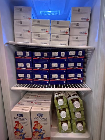 The image shows the interior of a refrigerator stocked with various milk products. The top shelf contains several white cartons with the word "МОЛОКО" (MILK) printed on them, along with dates and barcodes. The middle shelf is filled with blue cartons, also labeled "МОЛОКО" with a percentage indicating fat content (3.2%). The bottom shelf holds larger, white cartons featuring cartoon illustrations of people and the text "Хорошее дело МОЛОКО" (Good Deed MILK), again with a 3.2% fat indication and the word "ПРОСНУТЬСЯ И РАЗБУДИТЬ ВСЕХ" (Wake up and wake everyone up). To the right on the bottom shelf are green cartons labeled "planto BARISTA", indicating they are for coffee. The scene appears to be a domestic refrigerator. The location is identified as Veshki, Russia.