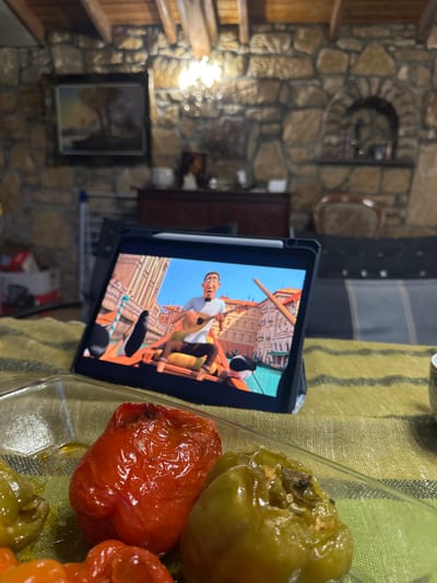 The image shows a casual indoor setting, likely a home. A tablet device displays a cartoon scene depicting a male character in what appears to be a gondola on a waterway, with architectural buildings in the background. The tablet rests on a surface covered with a striped green and yellow cloth. To the right of the tablet, there's a clear container holding what seems to be a selection of cooked bell peppers, varying in color from green to red. The background reveals a stone wall with embedded decorative elements, including a framed picture. The overall scene suggests a relaxed environment with the presence of technology, food, and unique architectural details. There is no indication of Pliska, Bulgaria.