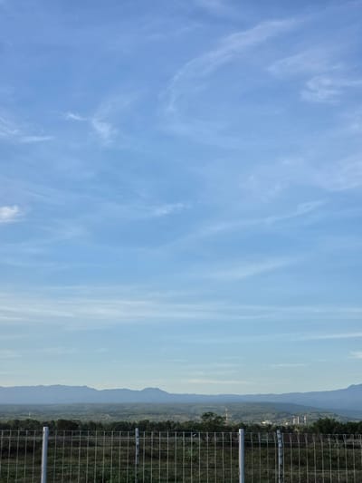 The image shows a view from Pintomakmur, Indonesia. A clear blue sky with wispy clouds occupies the left side of the frame. A mountain range is visible in the background, slightly obscured by haze. A wire fence runs along the right side of the image. A few buildings and vegetation are visible on the mountain side.