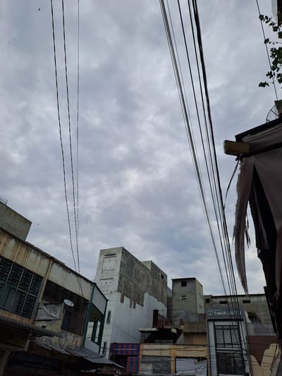 The image captures an overcast sky over a dense urban area, likely in Bandarbireuen, Indonesia, characterized by a clutter of overhead electrical wires and buildings. In the foreground and midground, several multi-story buildings are visible. The building on the far left has a weathered, aged facade with barred windows and a sign that appears to read "AQUEN PLASTIK." Adjacent to it is another building with a mix of white and grey concrete, showing signs of disrepair with patches of peeling paint and some windows. Further back, more structures rise, including one with a prominent sign for "APOTEK TIMUR LAUT 2." The overall impression is one of a bustling, possibly older, commercial district with a significant amount of exposed wiring contributing to a somewhat chaotic visual aesthetic. A few birds are present in the sky, and the general condition of the buildings suggests an environment that experiences significant weather exposure.