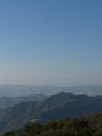 An elevated landscape photograph depicts a panoramic view of the Unincorporated Santa Monica Mountains, United States of America, under a clear, hazy blue sky. The upper two-thirds of the image features an unobstructed sky, transitioning from a deeper blue at the zenith to a lighter, more desaturated blue closer to the horizon line, indicative of atmospheric haze. The horizon reveals multiple undulating mountain ranges, progressively lighter and more indistinct with distance due to atmospheric effects. In the mid-ground, darker, vegetated mountain ridges are visible, displaying deep shadows on their slopes from a high-angle sun. Sparse, light-colored structures or developments are discernible within valleys and on distant hillsides. The immediate foreground shows the tops of dense, dark green scrub brush and chaparral, suggesting the viewpoint is from a vegetated elevated position. No individuals, animals, or specific human activities are present in the frame.